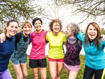 A line-up of six women standing outdoors dressed in exercise gear, smiling and with their arms around the person beside them.